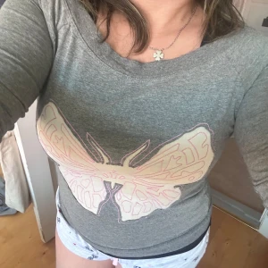 Grå långärmad tröja med fjärilstryck st s - Säljer en grå långärmad tröja från Delle med ett stort fjärilstryck i ljusa toner framtill. Tröjan har en bred rund halsringning och är perfekt för en avslappnad stil.