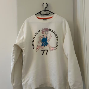 Vit sweatshirt Nike 'Honolulu Marathon '77' - Vit sweatshirt från Nike med tryck av en tecknad löpare och texten 'Honolulu Marathon '77' på framsidan. Stor Nike-logga på ryggen. Tillverkad i 100% bomull och har långa ärmar och rund hals.
