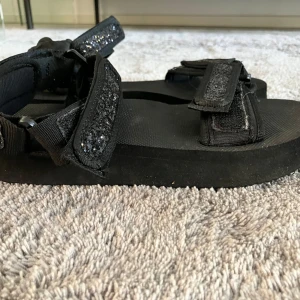 Svarta sandaler med glitterdetaljer - Säljer ett par svarta sandaler med justerbara kardborreband och glittriga detaljer på remmarna. Skorna har platt sula. Storlek 36/37