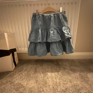 Blå volangkjol i jeans från Lindex - Supersöt blå jeanskjol med volanger från Lindex. Kjolen har resår i midjan och flera lager volanger som ger en riktigt fin och lekfull look. Perfekt för dig som gillar denim och vill ha något bekvämt men ändå trendigt.