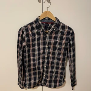 Rutig skjorta från Tommy Hilfiger - Snygg rutig skjorta från Tommy Hilfiger i mörkblått, vitt och rött. Klassisk krage och knäppning framtill. Perfekt för dig som gillar en tidlös och stilren look.