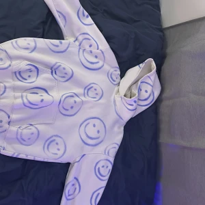 Vit hoodie med blå smiley-mönster - Säljer en vit hoodie med blåa smiley-ansikten över hela tröjan. Hoodien har en stor ficka framtill och en mysig huva. Perfekt för dig som gillar unika och lekfulla plagg!