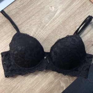 Svart spets-bh med rosett - Säljer en svart bh med vacker spets och liten rosett framtill. Bh:n har vadderade kupor och justerbara axelband. Perfekt för dig som vill ha något både snyggt och bekvämt. Knappast använd