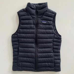 Svart dunväst från Patagonia - OANVÄND!!! Säljer en svart dunväst från Patagonia med quiltad design och dragkedja framtill. Västen har hög krage och klassisk passform, perfekt för lager-på-lager. Ikonisk Patagonia-logga på bröstet. Nypris 2499kr