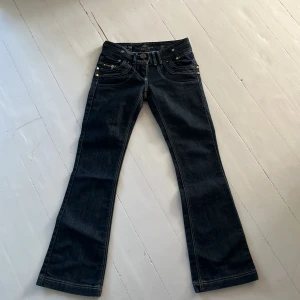 Mörkblå jeansbyxor från River Island med dragkedjedetaljer - Snygga mörkblå jeansbyxor från River Island med flera dragkedjor och dekorativa sömmar. Byxorna har bootcut-modell och låg midja, samt broderad logga på bakfickan. Perfekta för dig som gillar detaljer och unik stil. Midjemått rakt över 35cm, innerbenlängd 75cm.💗