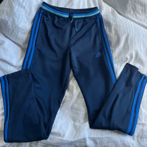 Mörkblå träningsbyxor från Adidas med blå ränder - Säljer ett par mörkblå träningsbyxor från Adidas med klassiska blå ränder längs sidorna och logga på låret. Byxorna har resår och dragsko i midjan samt smal passform.