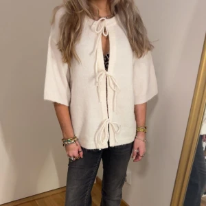 Vit kofta med knytband - Säljer en söt vit kofta med tre knytband framtill och kortare ärmar. Koftan är stickad och har en avslappnad passform, perfekt att slänga över en topp. Superfin detalj med knytningarna framtill!