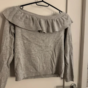 Ljusgrå tröja med volang - Säljer en ljusgrå långärmad tröja med bred volang runt axlarna. Tröjan har en offshoulder-design och är tillverkad i mjukt stickat material. Perfekt för dig som vill ha en trendig och bekväm look.