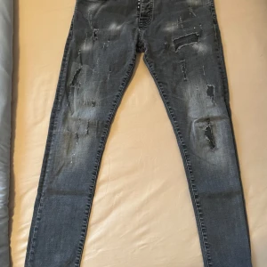 Grå slitna jeansbyxor DQ25 - Säljer ett par grå jeansbyxor från DQ25 med slitningar och distressed detaljer framtill. Byxorna har normal passform och klassisk femficksdesign. Perfekta för dig som gillar en tuff och avslappnad stil.