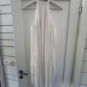 Vit plisserad klänning med halterneck - Säljer en elegant vit klänning med plisserat tyg och halterneck. Klänningen är ärmlös och har en luftig, flowy passform som ger en stilren och modern look. Perfekt för sommarens speciella tillfällen. Storleken står inte i men skulle säga S/M. 