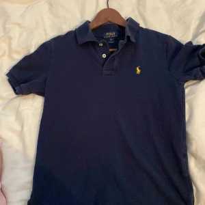 Mörkblå pikétröja från Polo Ralph Lauren - Klassisk mörkblå pikétröja från Polo Ralph Lauren med gul broderad logga på bröstet. Tröjan har korta ärmar, krage och knappar framtill. Perfekt för en stilren och avslappnad look. 