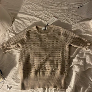 Beige stickad tröja från Holly & Whyte - Säljer en beige stickad tröja från Holly & Whyte med diskret svart randigt mönster. Tröjan har rund halsringning och ribbade muddar vid ärmslut och nederkant. Perfekt för en stilren och enkel look.