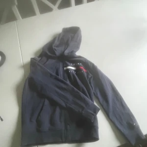 Mörkblå hoodie från Tommy Hilfiger - Säljer en mörkblå hoodie från Tommy Hilfiger med dragkedja och broderad logga på bröstet. Tröjan har långa ärmar och klassisk huva. Ikoniska ränder i vitt, rött och blått på bröstet samt liten logga på ärmen.