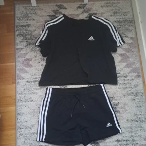 Svarta träningsshorts och t-shirt från Adidas - Svarta träningsshorts och t-shirt från Adidas med klassiska vita ränder längs sidorna och liten Adidas-logga framtill. Shortsen har elastisk midja med snörning och är tillverkade i ett mjukt syntetmaterial. Perfekta för sport eller gym. 