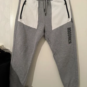 Grå mjukisbyxor från Hoodrich - Snygga grå mjukisbyxor från Hoodrich med vita paneler och svarta dragkedjor på fickorna. Byxorna har resår och snörning i midjan samt Hoodrich-logga på benet. Perfekta för en avslappnad stil.