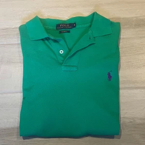 Grön pikétröja från Polo Ralph Lauren - Säljer en klassisk grön pikétröja från Polo Ralph Lauren i modellen Custom Fit. Tröjan har korta ärmar, krage och knäppning framtill samt den lila broderade loggan på bröstet.