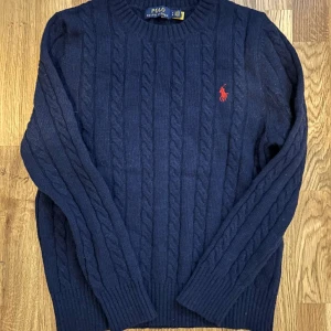 Mörkblå stickad tröja från Polo Ralph Lauren - Mörkblå stickad tröja från Polo Ralph Lauren med rund halsringning och röd broderad logga på bröstet. Klassisk modell med ribbade muddar vid ärmslut och nederkant. Vid funderingar, hör av er!