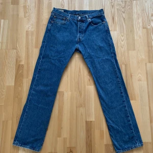 Levi's 501 blå jeans - Klassiska blå Levi's 501 jeans med raka ben och normal passform. Jeansen har fem fickor och är tillverkade i slitstarkt denim. Perfekta för dig som gillar tidlös stil och vill ha ett par ikoniska jeans i garderoben.