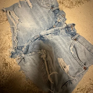 Ljusblå jeansshorts med slitningar från Zara - Säljer ett par ljusblå jeansshorts från Zara med råa kanter och slitna detaljer. Shortsen har medel midja, knappar framtill och klassiska fickor både fram och bak. Perfekta för en avslappnad stil. Sitter som en S-M🥰