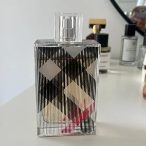 Burberry Brit Eau de Parfum - Burberry Brit Eau de Parfum i en rektangulär glasflaska med det klassiska rutmönstret i svart, vitt och rött. Flaskan har en transparent spraykork och stilren design som känns både modern och tidlös. Perfekt för dig som gillar eleganta och ikoniska dofter. Nästan 100ml