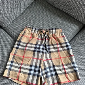 Rutig shorts från Burberry - Säljer ett par klassiska Burberry shorts med det ikoniska rutmönstret i beige, svart, vitt och rött. Shortsen har resår i midjan och snörning för justerbar passform. Perfekta för sommaren och riktigt snygga till en enkel t-shirt.