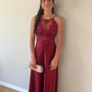 Vinröd långklänning med spets och satin - Vinröd långklänning med spetsdetaljer upptill och smala axelband. Klänningen har en elegant v-ringning med mesh och omlottkänsla i spetsen, samt en slät satinkjol. Perfekt för bal eller festliga tillfällen.