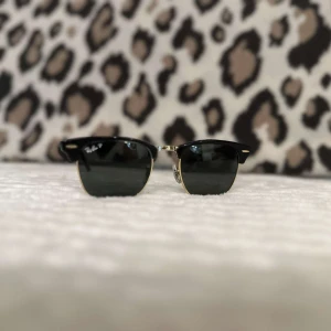 Svarta solglasögon från Ray-Ban - Klassiska svarta solglasögon från Ray-Ban med halvbåge och mörka glas. Metallbåge i guldton och svarta detaljer upptill. Kommer med original svart fodral i skinnimitation.