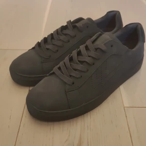 Mörk Gråa lejon sneakers från skopunkten - Säljer ett par mörk gråa sneakers från skopunkten med klassisk design och rund tå. Skorna har snörning och är tillverkade i syntetmaterial. Perfekta för en stilren och enkel look. Har aldrig använt dem