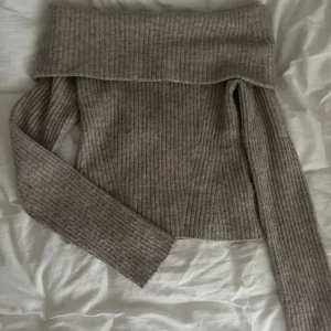 Grå ribbad offshoulder stickad tröja från H&M - Säljer en grå stickad tröja från H&M med ribbat material och offshoulder-design. Tröjan har långa ärmar och en bred kant upptill som viks ner över axlarna. Perfekt för en trendig och mysig look.💕