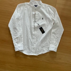 Vit skjorta från Ralph Lauren - Klassisk vit skjorta från Ralph Lauren med broderad logga på bröstet. Skjortan har lång ärm, knappar framtill och är tillverkad i bomull. Perfekt för en stilren och tidlös look.
