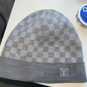 Grå rutig mössa från Louis Vuitton - Snygg grå mössa från Louis Vuitton med klassiskt rutigt mönster och LV-logga framtill. Mjuk och bekväm passform, perfekt för kyliga dagar. Diskret men lyxig accessoar som lyfter din outfit.