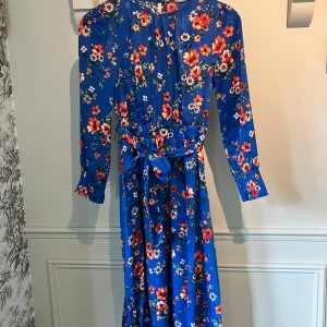 Blommig blå långklänning från Orsay - Säljer en blå långklänning från Orsay med färgglatt blommönster. Klänningen har långa ärmar, knytband i midjan och rund halsringning. Perfekt för dig som vill sticka ut med en färgstark och feminin look.