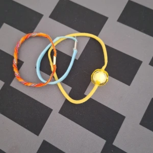 Färgglada armband i olika material - Tre armband i olika färger och material. Ett är orange och flerfärgat i tråd, ett är ljusblått och ett är gult med en rund gul pärla. Perfekta för att addera färg till din stil. Säljer för 5kr.