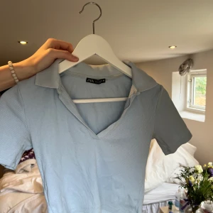 Ljusblå croppad från Zara - Säljer en ljusblå croppad från Zara med korta ärmar och v-ringning. Toppen har en klassisk krage och är tillverkad i ett mjukt material som känns bekvämt mot huden. Storlek S