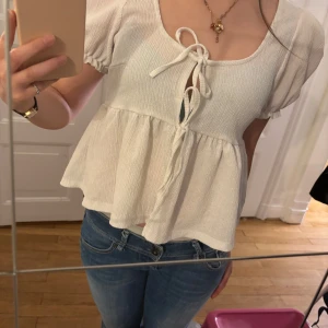Vit blus med puffärm och knyt från Gina Tricot - Supersöt vit blus från Gina Tricot med kort puffärm och två knyt framtill. Blusen har en lätt rynkad, volangig nederdel och är tillverkad i ett strukturerat bomullsliknande material. Perfekt för en somrig look!
