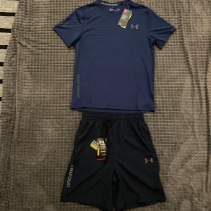 Under Armour sett  - Snygga svarta träningsshorts från Under Armour med elastisk midja och snörning. Shortsen har logga på ena benet och texten 'ARMOUR' i färg längs sidan. Perfekta för gym eller löpning.