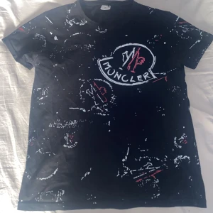 Svart monclare t shirt - Svart t-shirt från Moncler med vitt och rött tryck och ett stort Moncler-logo på bröstet. T-shirten har korta ärmar och ett unikt, abstrakt mönster över hela plagget. Perfekt för dig som gillar streetwear och vill sticka ut.
