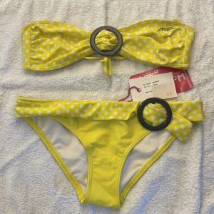 Gul prickig bikini från Protest - Säljer en söt gul bikini från Protest med vita prickar och gråa ringdetaljer. Överdelen är bandeau-modell med knyt i ryggen och underdelen har en dekorativ rosett framtill. Perfekt för stranden eller poolen! Bikinin är helt oanvänd med trosskydd och prislappar kvar! Pris kan diskuteras. 