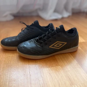 Svarta fotbollsskor från Umbro - Säljer ett par svarta fotbollsskor från Umbro med guldiga detaljer och logga på sidan. Skorna har snörning och en låg profil med platt sula, perfekt för inomhusspel. Snyggt mönster på ovandelen och klassisk design.
