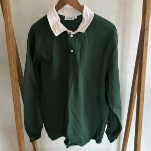 Grön pikétröja med vit krage från Weekday - Säljer en grön långärmad pikétröja från Weekday med klassisk vit krage och knäppning framtill. Perfekt för en stilren och avslappnad look. Passar dig som gillar enkel men snygg design.