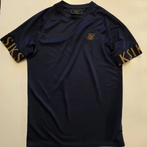 Mörkblå t-shirt från SikSilk med gulddetaljer - Snygg mörkblå t-shirt från SikSilk. Tillverkad i ett mjukt och lätt syntetmaterial – perfekt för sommaren. En av de bästa t-shirts jag någonsin har ägt! Sitter tajt över biceps och ger en riktigt bra "pump"-effekt, samtidigt som den är jätteskön att ha på sig. Tyvärr blev just den här lite för liten för mig, men kommer troligtvis köpa en ny i större storlek. T-shirten är i perfekt skick och märks inte att den är använd.  STORLEK M  ord. Pris: 599 kr