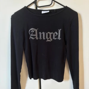 Svart långärmad topp med strass-text - Svart långärmad topp från STAY med texten 'Angel' i strass framtill. Tight passform och rund halsringning. Perfekt för dig som gillar statement-plagg med lite edge.