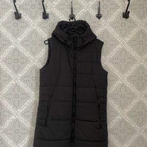 Svart lång dunväst från Jack Wolfskin - Svart lång dunväst från Jack Wolfskin med huva. Har använt ett fåtal gånger.