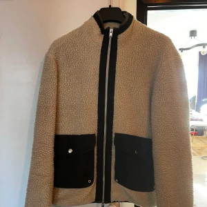Selected Homme – Beige jacka i teddy med kontrastdetaljer - Säljer en beige teddyjacka med svarta fickor och dragkedja framtill. Jackan har hög krage och två stora fickor med lock och knapp. Perfekt för kyliga dagar när du vill vara både varm och snygg.En väldigt snygg jacka som är använd ett fåtal gånger  https://www.asos.com/se/selected-homme/selected-homme-beige-jacka-i-teddy-med-kontrastdetaljer/prd/200842221