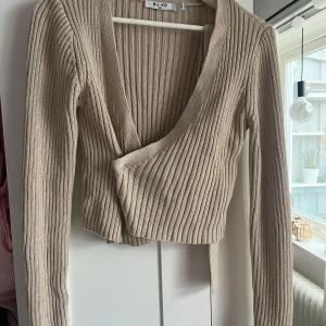 Beige ribbad omlottkofta från NA-KD - Trendig beige ribbad omlottkofta från NA-KD med långa ärmar och knyt framtill. Croppad modell som passar perfekt till både jeans och kjol. Mjuk stickad kvalitet och snygg passform. Knappt använd. Skriv för fler bilder💞