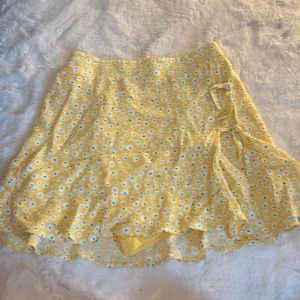 Gul blommig kjol med volang från Zara - Supersöt gul kjol från Zara med små vita och blå blommor. Kjolen har volangkant och omlottkänsla med knyt i sidan. Inbyggda shorts under för extra komfort, passar mig som är 164cm!