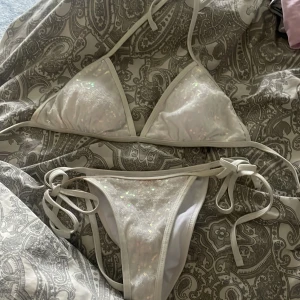 Vit glittrig bikini med paljetter - Säljer en snygg vit bikini med glittriga paljetter och knyt i sidorna och nacken. Bikinin har klassisk triangelmodell upptill och smala band. Perfekt för sommaren och strandhäng!