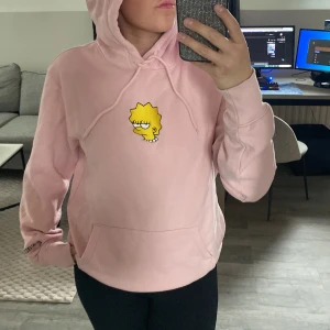 Rosa hoodie med Lisa Simpson-tryck - Säljer en ljusrosa hoodie med tryck av Lisa Simpson på bröstet. Tröjan har huva med dragsko och en stor magficka framtill. 
