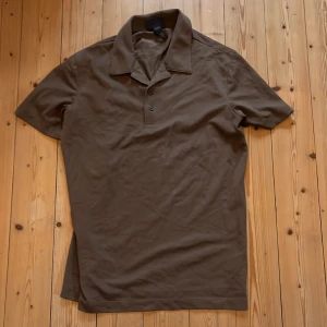 Brun piké t-shirt från H&M - Säljer en brun piké t-shirt från H&M i storlek XS. T-shirten har korta ärmar, klassisk krage och knäppning framtill. Perfekt för en avslappnad och stilren look.