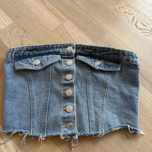 Jeans tubtopp med knappar - Säljer en cool tubtopp i jeans med knappar framtill och två dekorativa fickor. Toppen har råa, fransiga kanter och smock baktill för bättre passform. Perfekt för dig som gillar denim och vill sticka ut!